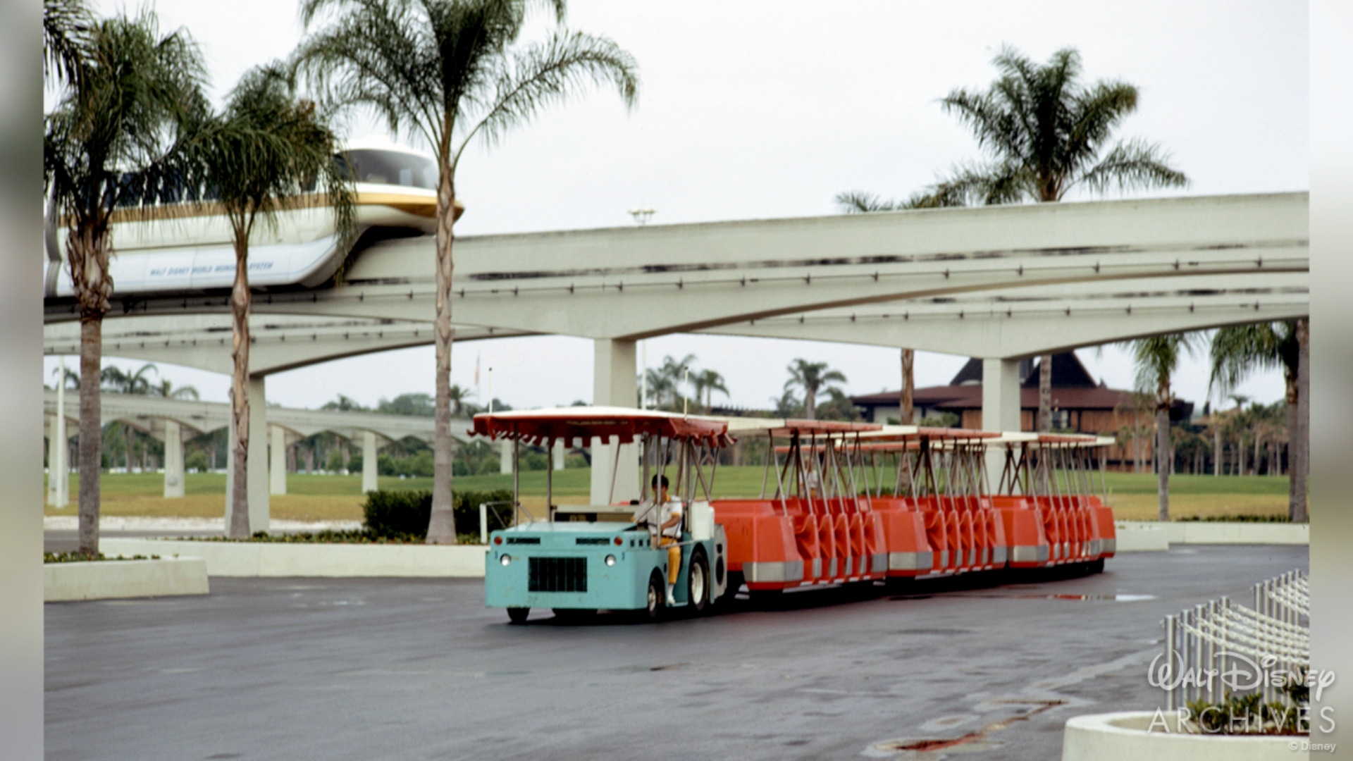 A Monorail with a gold stripe is positioned on the elevated track in the upper left corner of the image. Beneath the track, on a main road, is a parking tram used to transport guests to and from their cars. The driver&rsquo;s car is a robin&rsquo;s egg blue, and the passenger cars are a deep pastel red. 