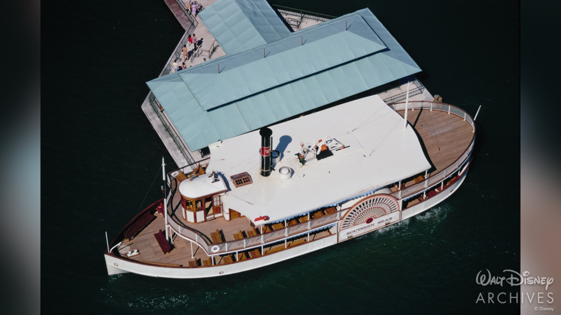 An aerial view of the Southern Seas water vessel at a dock. It is two story, medium-sized white ship. The decks of the vessel are wooden, and the dock&rsquo;s awning is blue. 