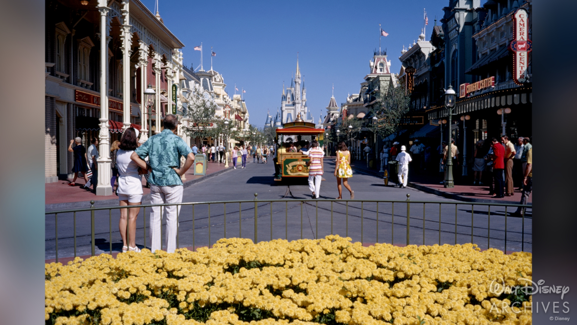 Photo taken from the middle of Town Square, a small hub at the entrance of Main Street, U.S.A., allowing for a straight view of nearby shops and Cinderella Castle. Small yellow flowers bloom in the foreground. Two guests stand with their backs to the camera and admire Cinderella Castle and the Main Street Trolley. Other guests mill about the street in the background. 