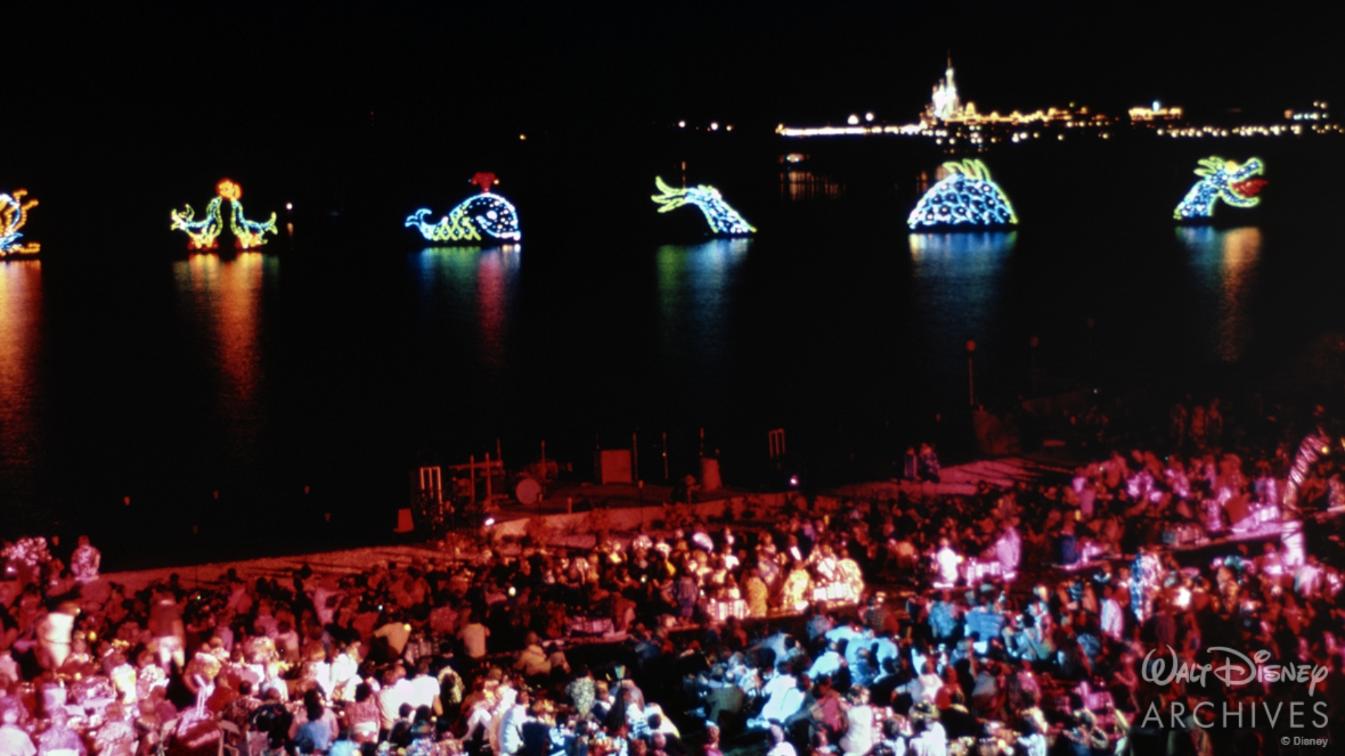 Guests watch a nighttime spectacle, the &ldquo;Electrical Water Pageant,&rdquo; from tables filling the beach of Disney&rsquo;s Polynesian Village Resort, along the Seven Seas Lagoon. Illuminated lights on a series of barges create the colorful shapes of a serpent, whale, and seal over the lagoon. In the distance, Cinderella Castle illuminates the sky. 