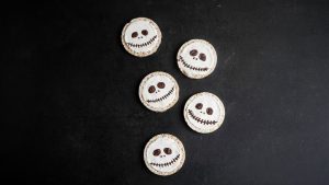 Jack Skellington-inspired rice cakes decorated with white frosting and chocolate facial features are arranged on a dark surface for a The Nightmare Before Christmas recipe.