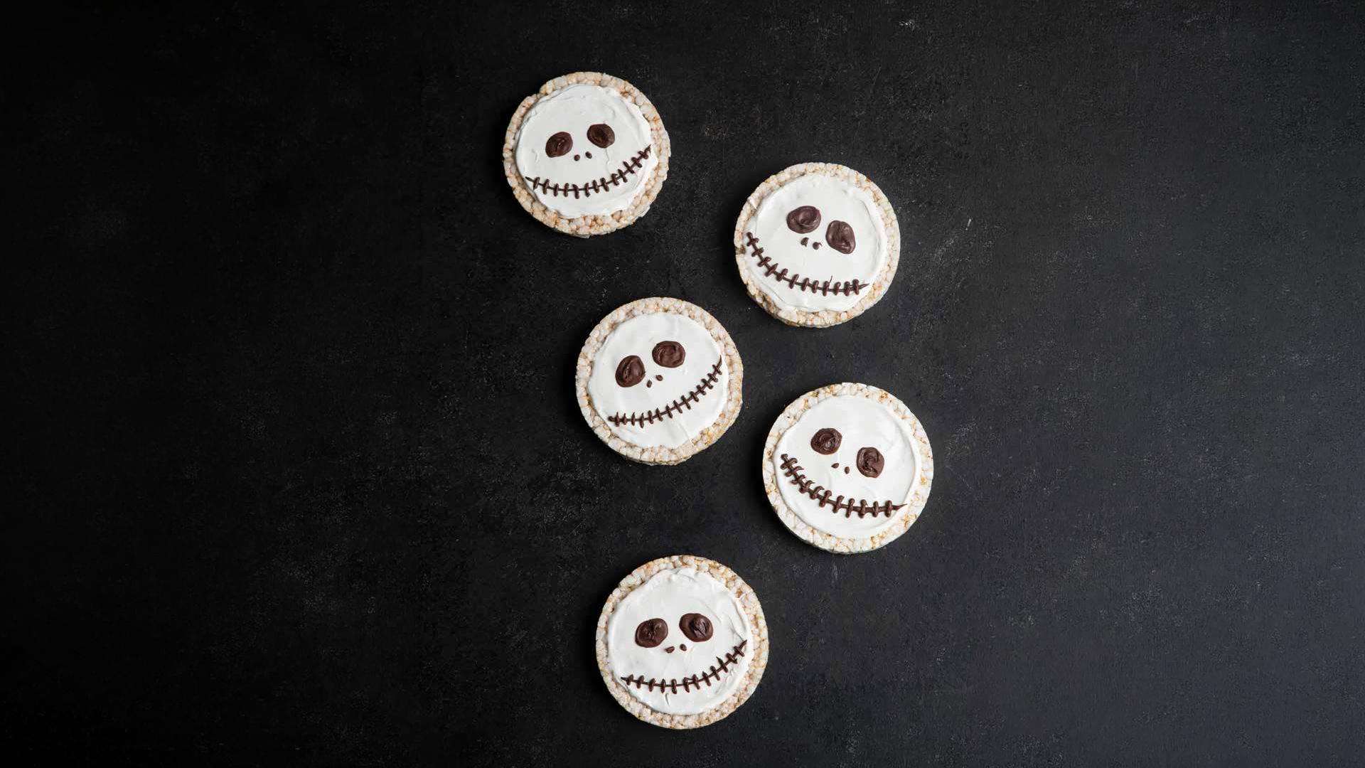Jack Skellington-inspired rice cakes decorated with white frosting and chocolate facial features are arranged on a dark surface for a The Nightmare Before Christmas recipe.