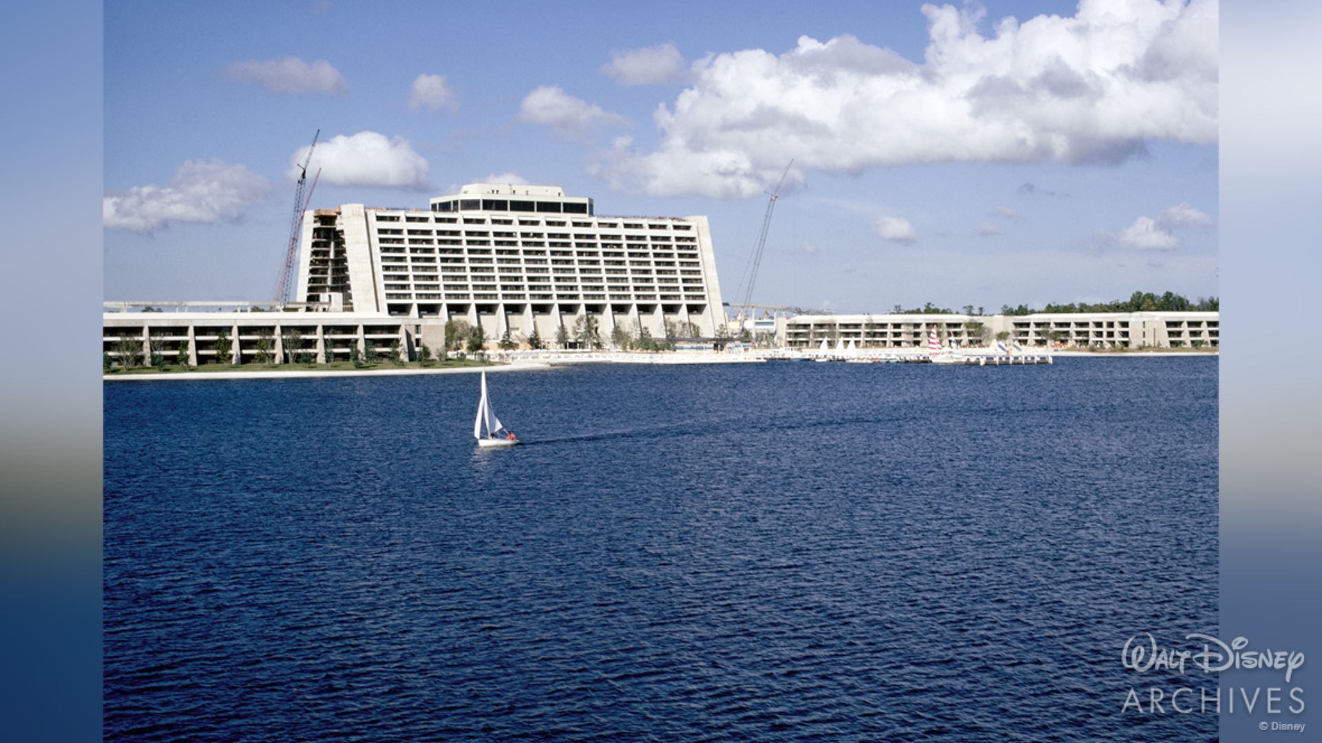 The white concrete A-Frame of Disney&rsquo;s Contemporary Resort looms over the deep blue Bay Lake. The image is taken from the back of the of hotel, featuring the marina, which is home to several boats is in view. A single sailboat is gliding through the lake. 