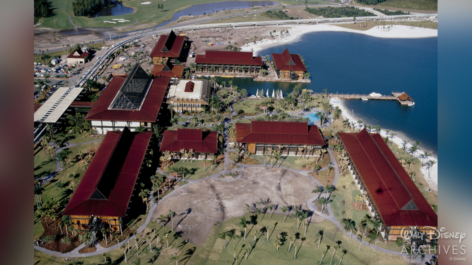 An aerial view of Disney&rsquo;s Polynesian Village Resort, featuring all eight longhouses, the Great Ceremonial House, the marina, pool, and Monorail station. The resort&rsquo;s longhouses are enhanced by high‐peaked, rust‐colored roofs. 