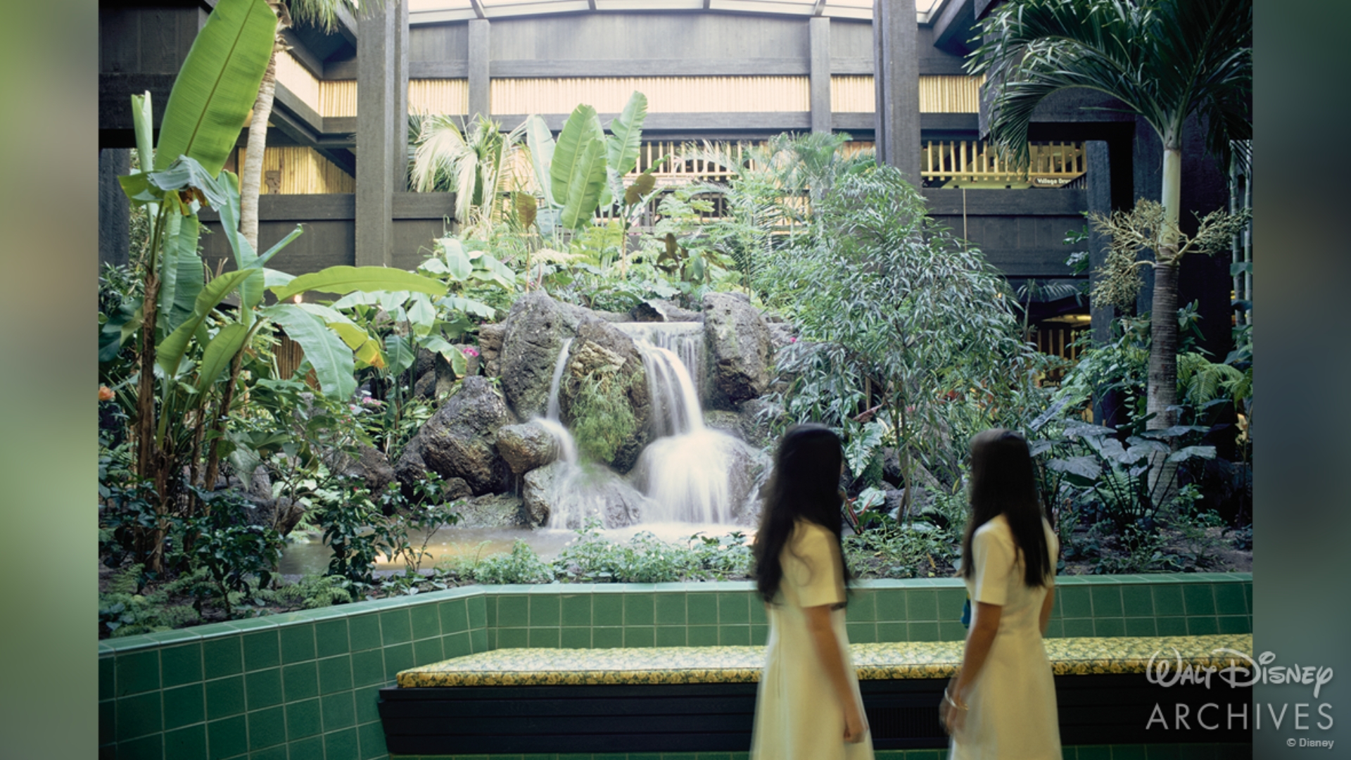 The centerpiece of the lobby features a leafy green fauna installation that reaches to the second floor. In the middle, a rocky structure with running water creates a small waterfall. Two young guests, wearing white dresses, are facing away from the camera towards the small garden. 
