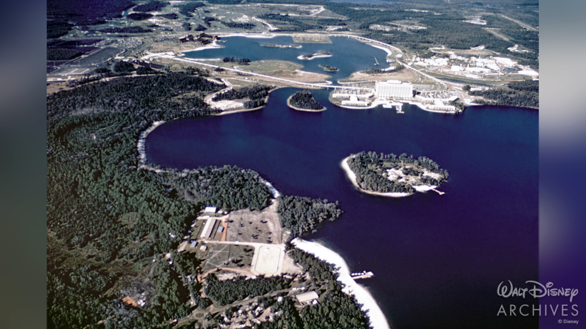 A wide shot of the Walt Disney World property in 1971. Toward the bottom of the image is the Fort Wilderness campground on the shores of Bay Lake surrounded by a dense forest. The top of the photo shows more of the Disney property. The Contemporary and Polynesian Village resorts are visible as well as Magic Kingdom Park and connecting Monorail loop. 
