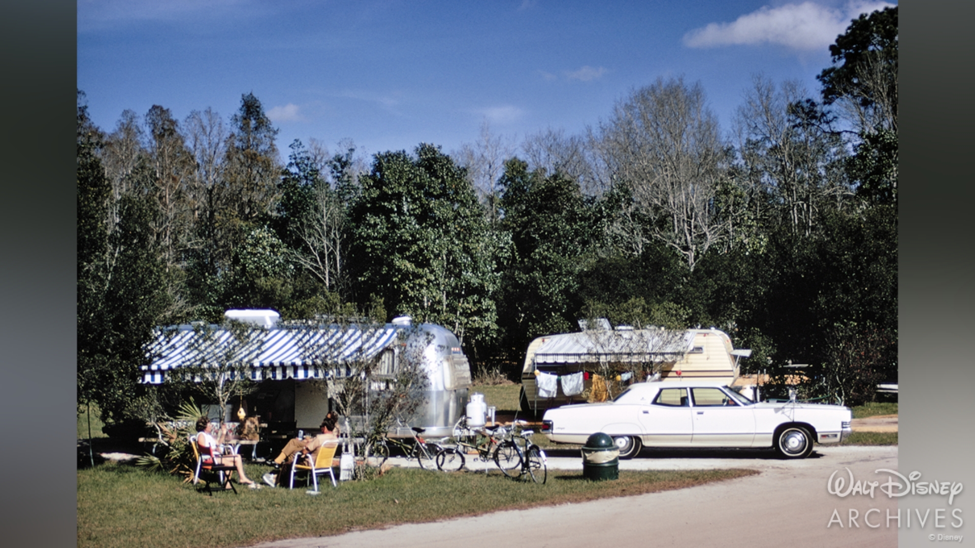 In front of a dense forest of trees, a silver Airstream sits hooked up to a white four door sedan at a Fort Wilderness campsite. A family relaxes in front of the Airstream in yellow lawn chairs with its bikes causally propped up on the grass. Another trailer can be seen at the next campsite. 