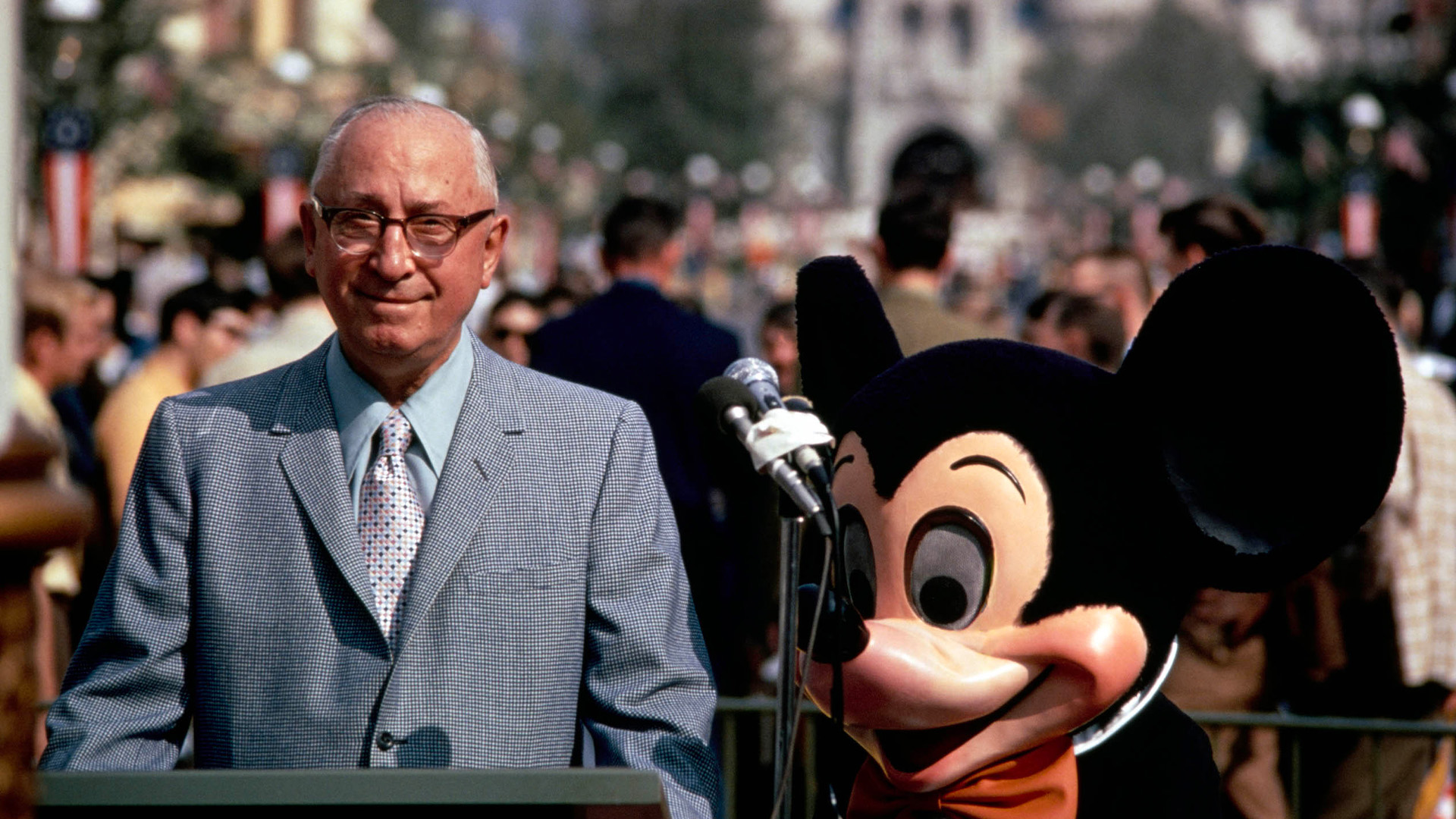 Roy O. Disney stands beside Mickey Mouse with the Walt Disney World dedication plaque in Town Square on Main Street, U.S.A. in the Magic Kingdom. Guests gather in the background, while a set of microphones stands between Roy and Mickey. Cinderella Castle appears softly out of focus in the background.