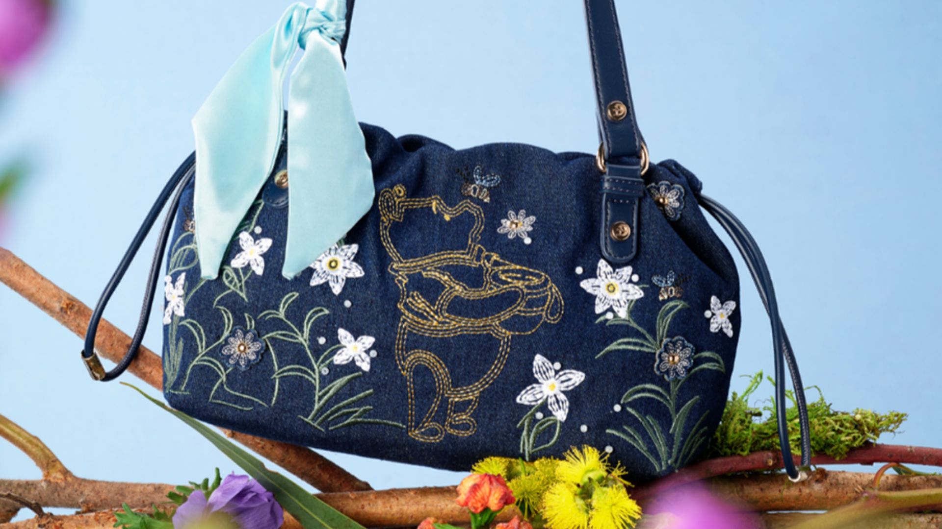 The Winnie the Pooh denim shoulder bag sits on a tree branch in front of a blue background. The stitching of the bag includes a yellow Winnie the Pooh holding his signature honey pot. Surrounding him are stitching&rsquo;s of flowers and bumble bees.
