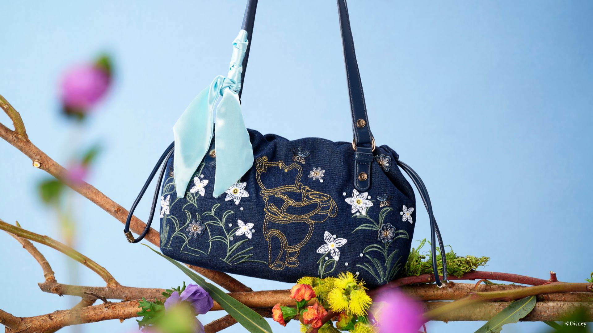 The Winnie the Pooh denim shoulder bag sits on a tree branch in front of a blue background. The stitching of the bag includes a yellow Winnie the Pooh holding his signature honey pot. Surrounding him are stitching&rsquo;s of flowers and bumble bees. 