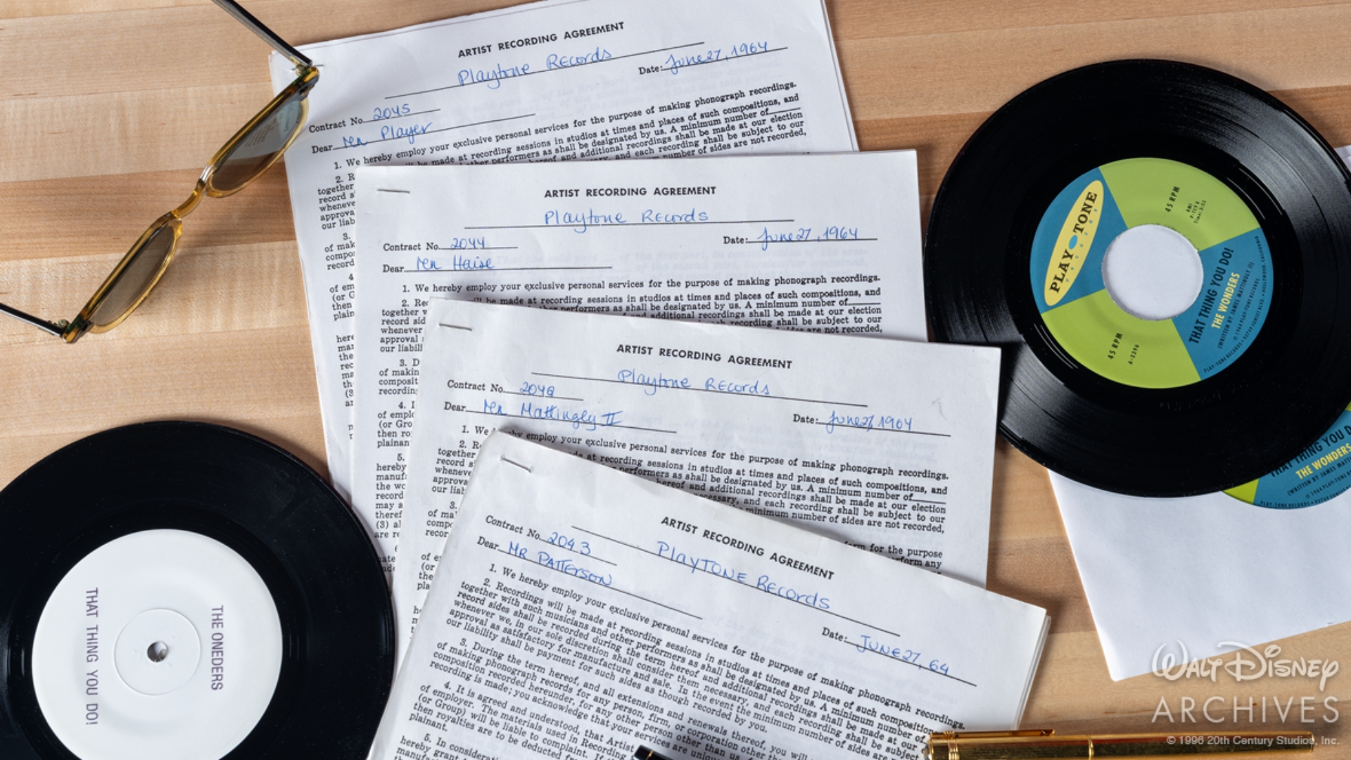 A collection of prop documents and records from That Thing You Do! is arranged on a wooden tabletop. Four Play-Tone Records artist recording agreements, dated June 27, 1964, are stacked in the center. Surrounding the contracts are two loose vinyl records, one labeled &ldquo;That Thing You Do!&rdquo; by the Wonders and another bearing the band&rsquo;s original name, the Oneders. A pair of gold-framed sunglasses, a gold pen, and another sleeved record complete the display.