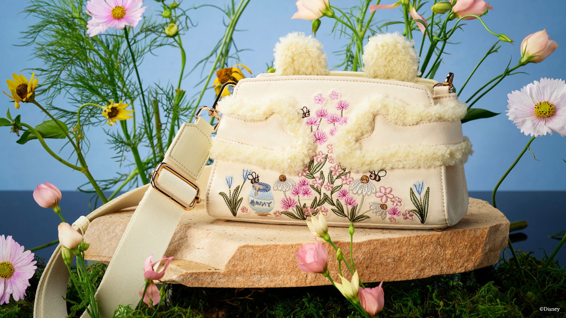 The Winnie the Pooh inspired Soft Embrace Satchel sits in front of a blue background surrounded by flowers. This softly textured handbag, pairs suedette with fluffy sherpa trim and Pooh ears peeking above the bag, while his cozy arms wrap around the front. Delicate embroidery featuring flowers, buzzing bees, and a hunny pot capture the whimsy of the Hundred Acre Wood against a soft pastel yellow suedette. A removable crossbody strap lets the wearer switch from satchel styling to hands-free wear, making this Winnie the Pooh handbag a charming companion for everyday outings and Disney days.