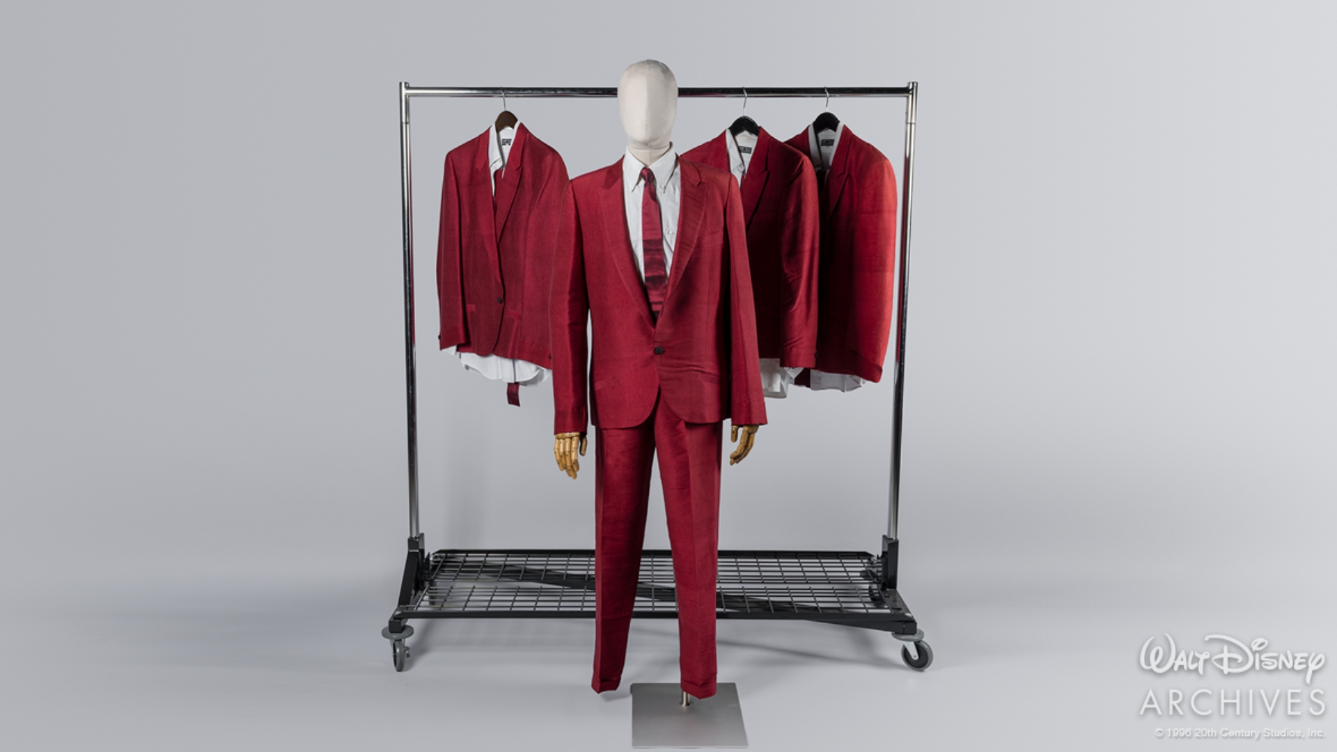 Four matching deep-red stage suits from That Thing You Do! are displayed on a rolling costume rack against a neutral gray background. In the foreground, Guy Patterson&rsquo;s costume is mounted on a mannequin and includes a red jacket and trousers, white dress shirt, and narrow red tie. Three additional band costumes hang behind it on the rack.