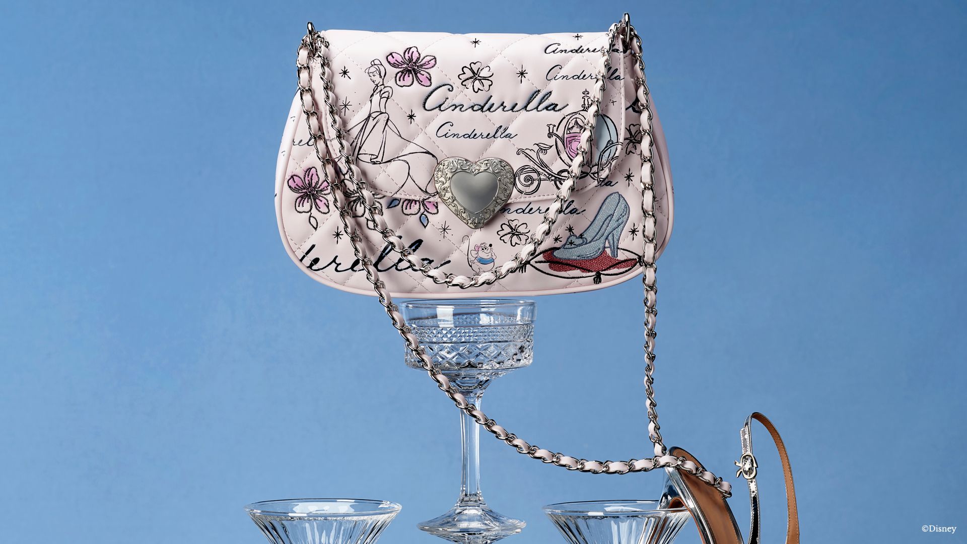 A light pink Cinderella inspired bag sits on top of an empty cocktail glass. Images on the bag include an outline of Cinderella, the pumpkin carriage, the glass iconic slipper and flowers. 