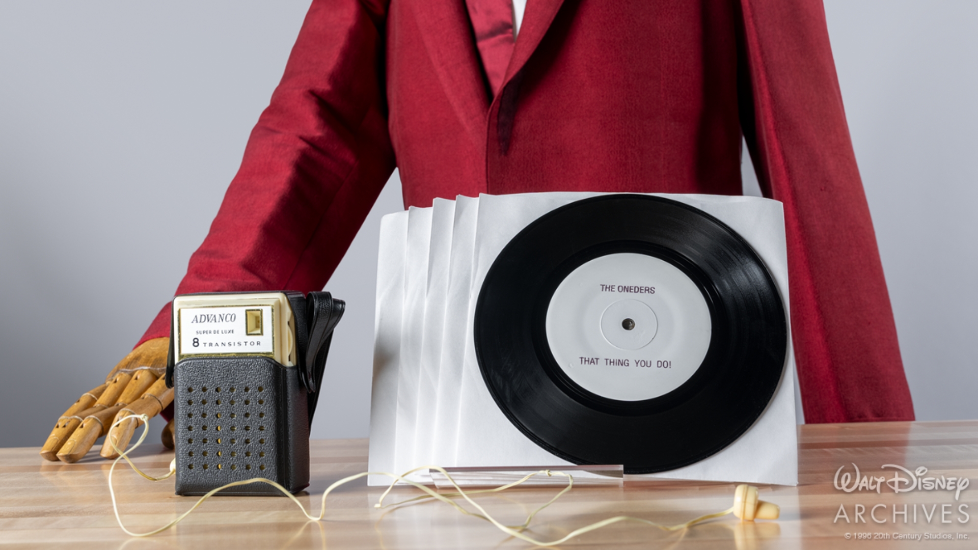 A display of props from That Thing You Do! featuring several vinyl singles and a vintage transistor radio. In the foreground, a black-and-silver portable radio with wired earbuds sits beside a stack of white record sleeves. A vinyl record is propped in front of a red stage suit worn by a member of the Wonders. The record&rsquo;s center label reads &ldquo;The Oneders&rdquo; above &ldquo;That Thing You Do!&rdquo; 