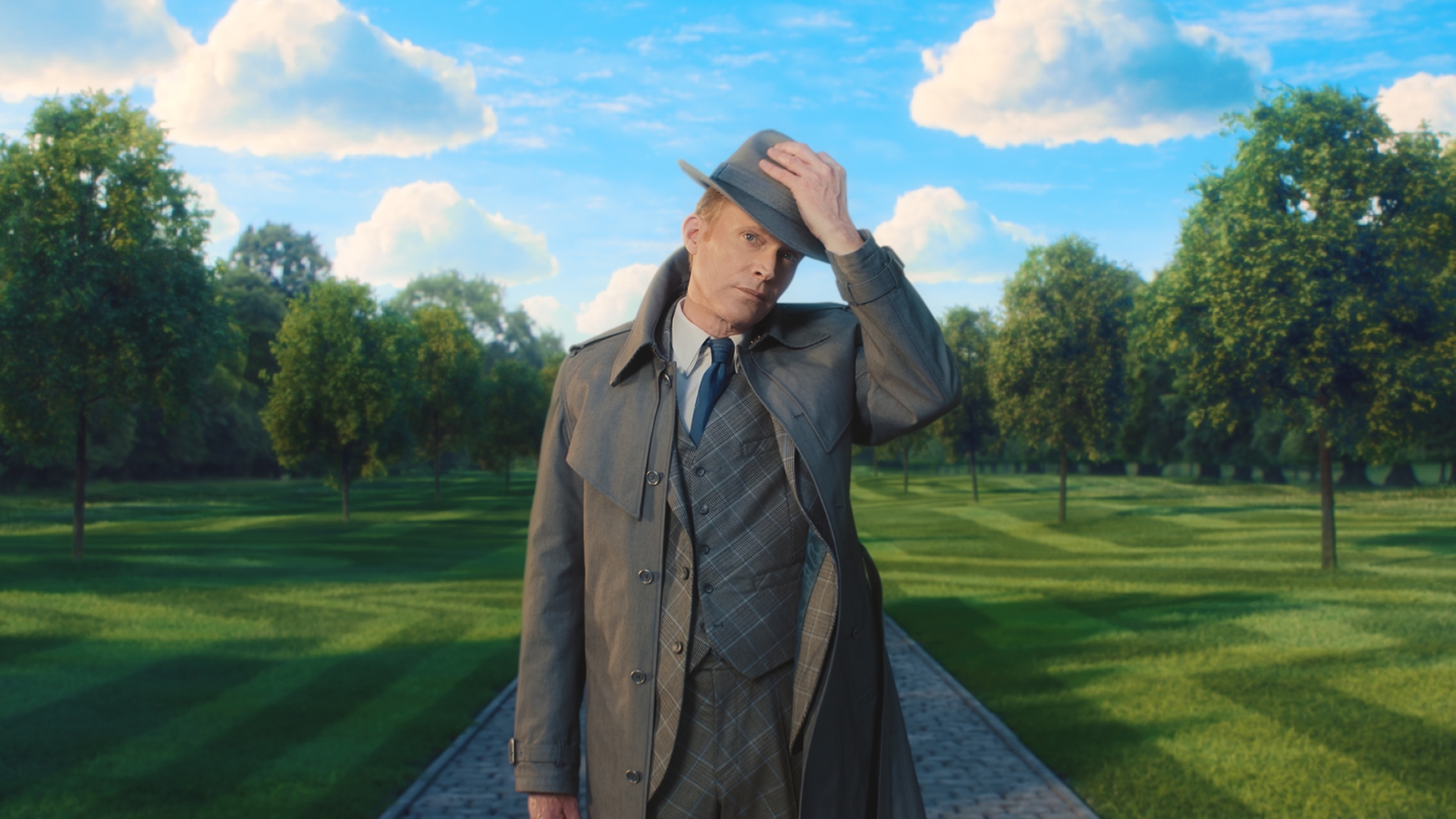 In this scene from Marvel Television&rsquo;s VisionQuest, Vision, played by Paul Bettany, appears in his human form and tips his hat. He is wearing a trench coat and walking on a path, surrounded by perfectly manicured green grass. Lush trees appear in the background.