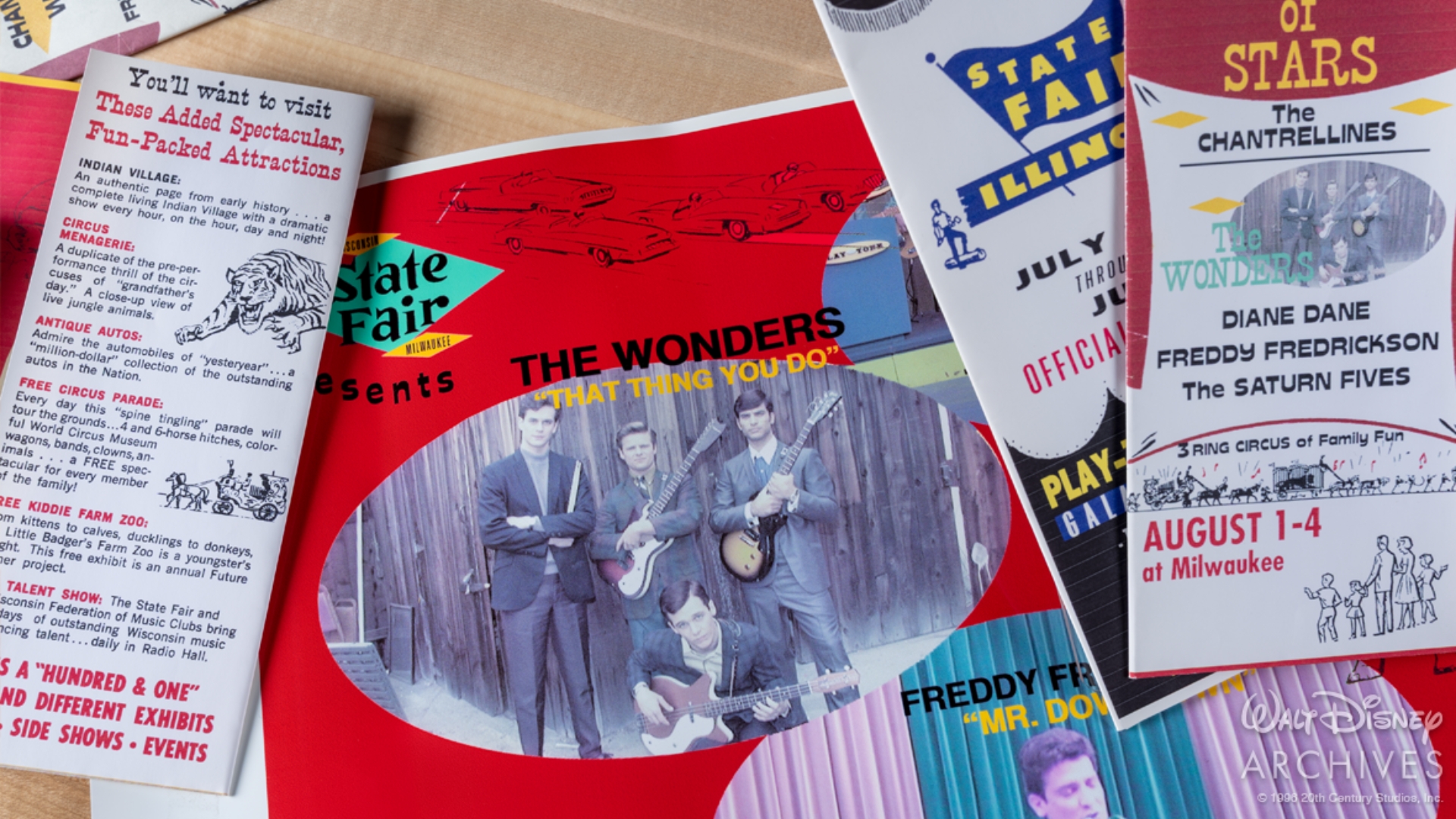 A collection of colorful prop promotional materials from the Play-Tone Galaxy of Stars Tour featured in That Thing You Do! is spread across a tabletop. At the center is a large state fair poster highlighting the Wonders and their hit song &ldquo;That Thing You Do!&rdquo; Surrounding the poster are prop brochures and tour advertisements listing performers including the Wonders, the Chantrellines, Diane Dane, Freddy Fredrickson, and the Saturn 5s. The materials are designed in an early-to-mid-1960s style with bright colors, illustrated fair attractions, and period typography.