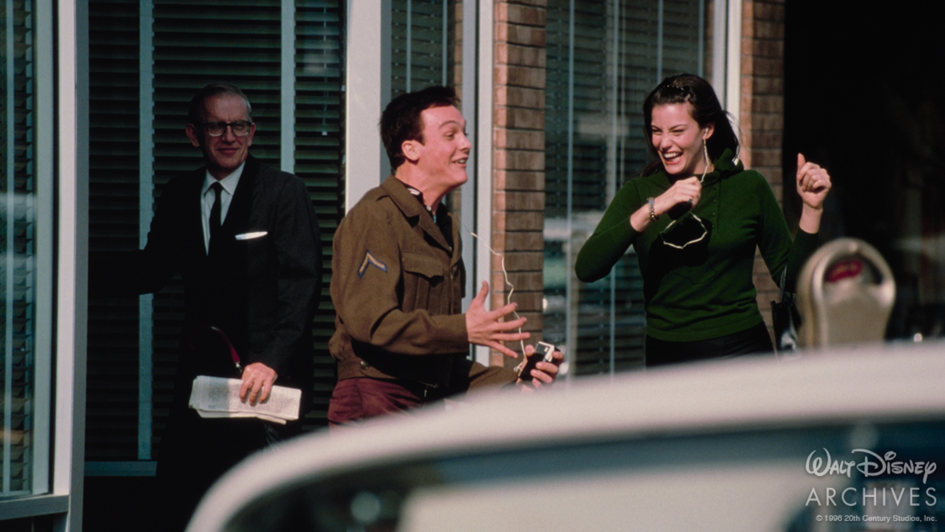 A production still from That Thing You Do! shows The Bass Player carrying a small transistor radio with an earbud cord trailing from it as he runs along a downtown sidewalk. Beside him, Faye Dolan keeps pace through a crowd of pedestrians. The scene is set on a busy commercial street with a parking meter set in the foreground.