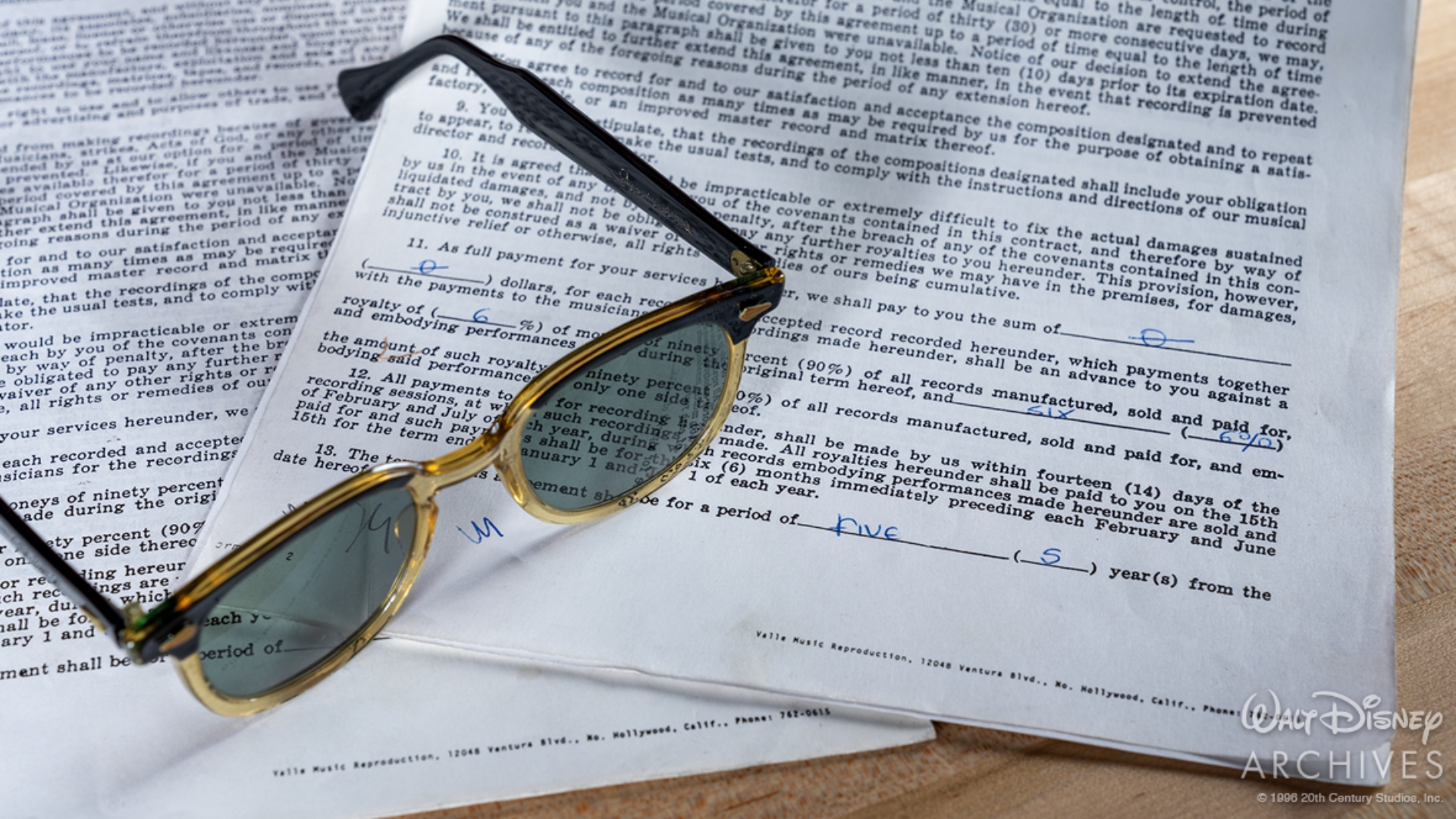 A pair of prop black-and-gold-framed sunglasses rests atop a stack of prop Play-Tone Records artist contracts on a wooden tabletop. The documents feature typed contract language with several handwritten markings. 