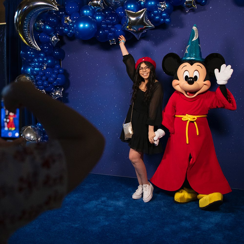 A female D23 Member poses with Sorcerer's Apprentice Mickey Mouse in front of a starry sky back drop.