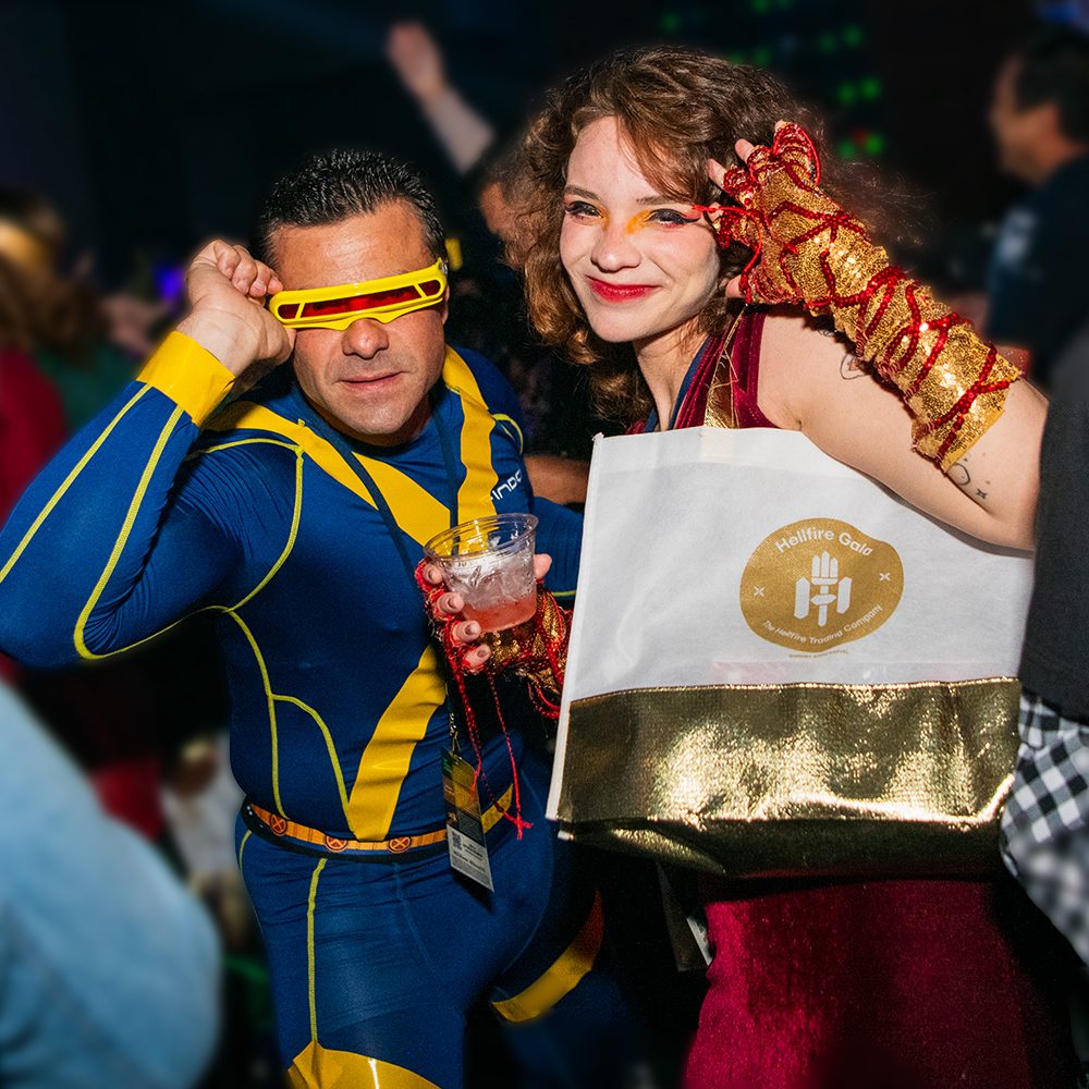 Two attendees at D23's Hellfire Gala in 2023. On the left, the attendee is dressed as Cyclops from Marvel's X-Men, complete with blue and yellow jumpsuit and yellow visor, which they are holding with one hand. On the right, the attendee has long red hair and is dressed in a red dress that is partially blocked by a white and gold Hellfire Gala swag bag hanging from their shoulder; they are holding up their red and gold-gloved hand to their face and smiling at the camera.