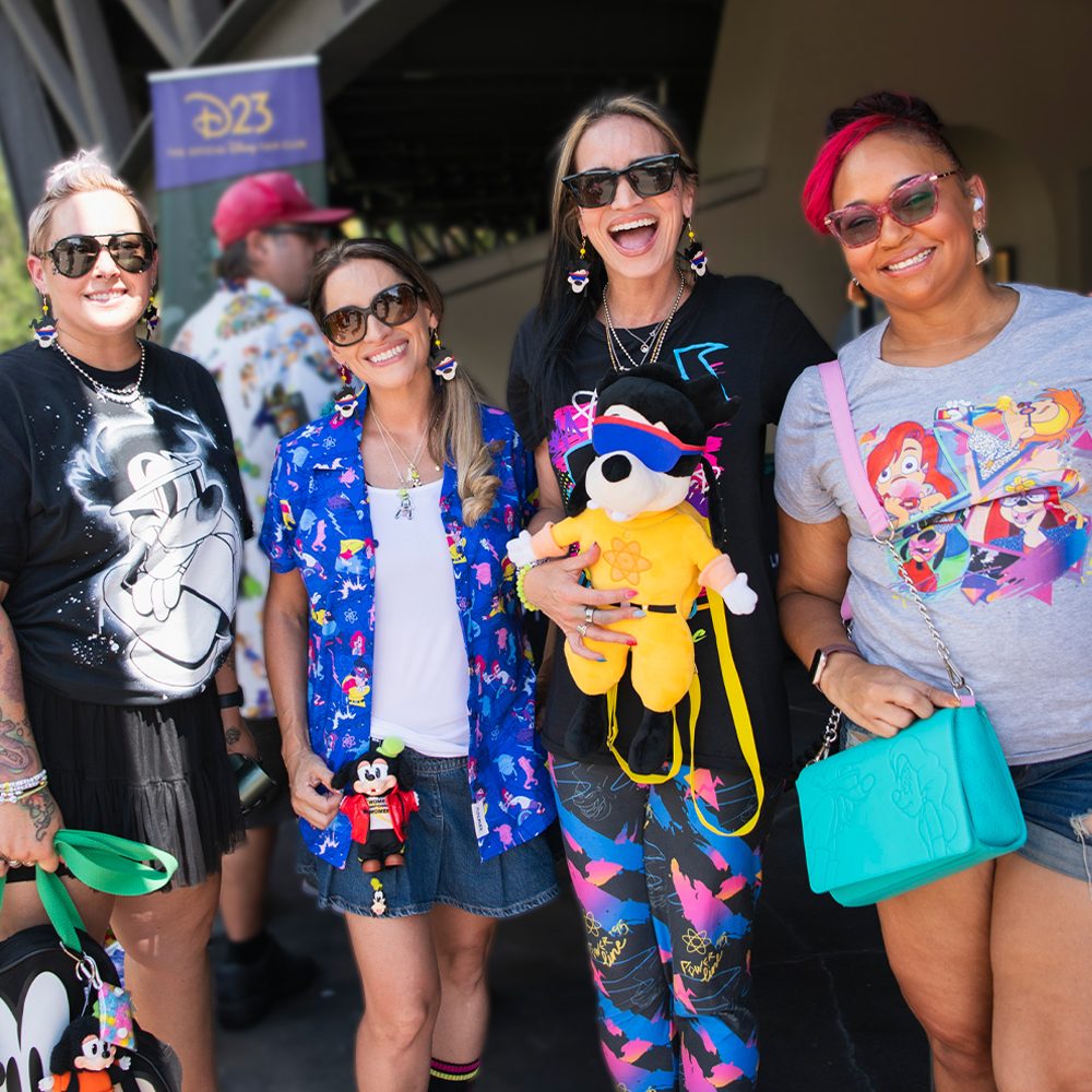 D23 Homepage Fans A group of four smiling Disney fans pose together at a D23 event, wearing colorful Disney-themed apparel including shirts featuring Goofy, Ariel, and other characters. One fan holds a plush of Max dressed in bright yellow, while another has a small Mickey plush attached to her outfit. All are accessorized with sunglasses and themed jewelry, standing in front of a D23 banner.