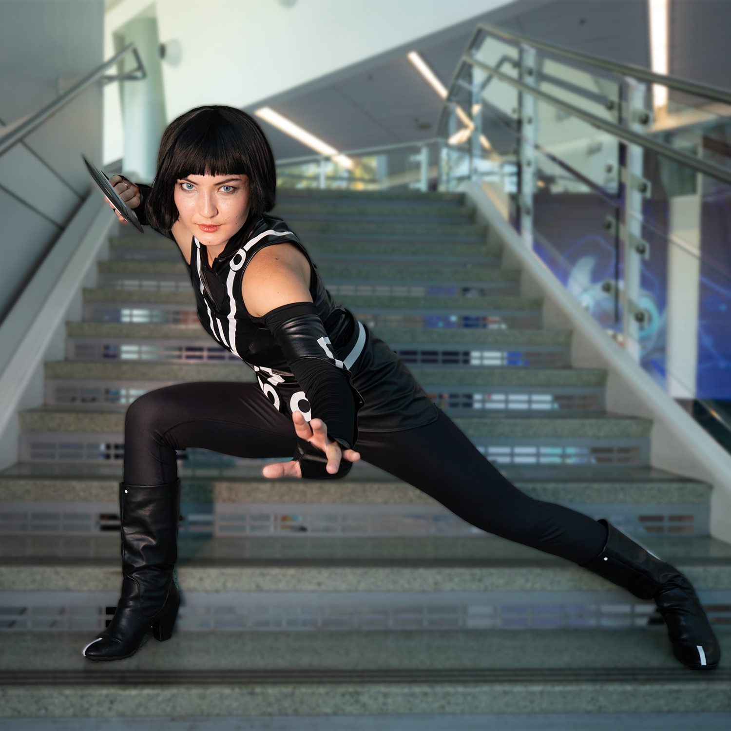 A cosplayer dressed in a sleek black TRON-inspired outfit crouches in a dynamic pose on a staircase, holding a glowing identity disc and staring intently forward.