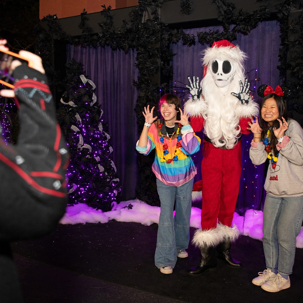 Two guests pose with Jack Skellington dressed as Sandy Claws at a festive photo spot featuring purple lights, dark holiday trees, and faux snow, with another person taking their picture in the foreground.
