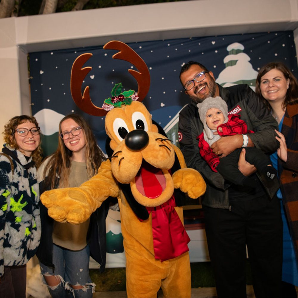 A family poses with Pluto dressed in festive reindeer antlers and a red holiday bow; everyone smiles brightly in front of a snowy winter-themed backdrop.