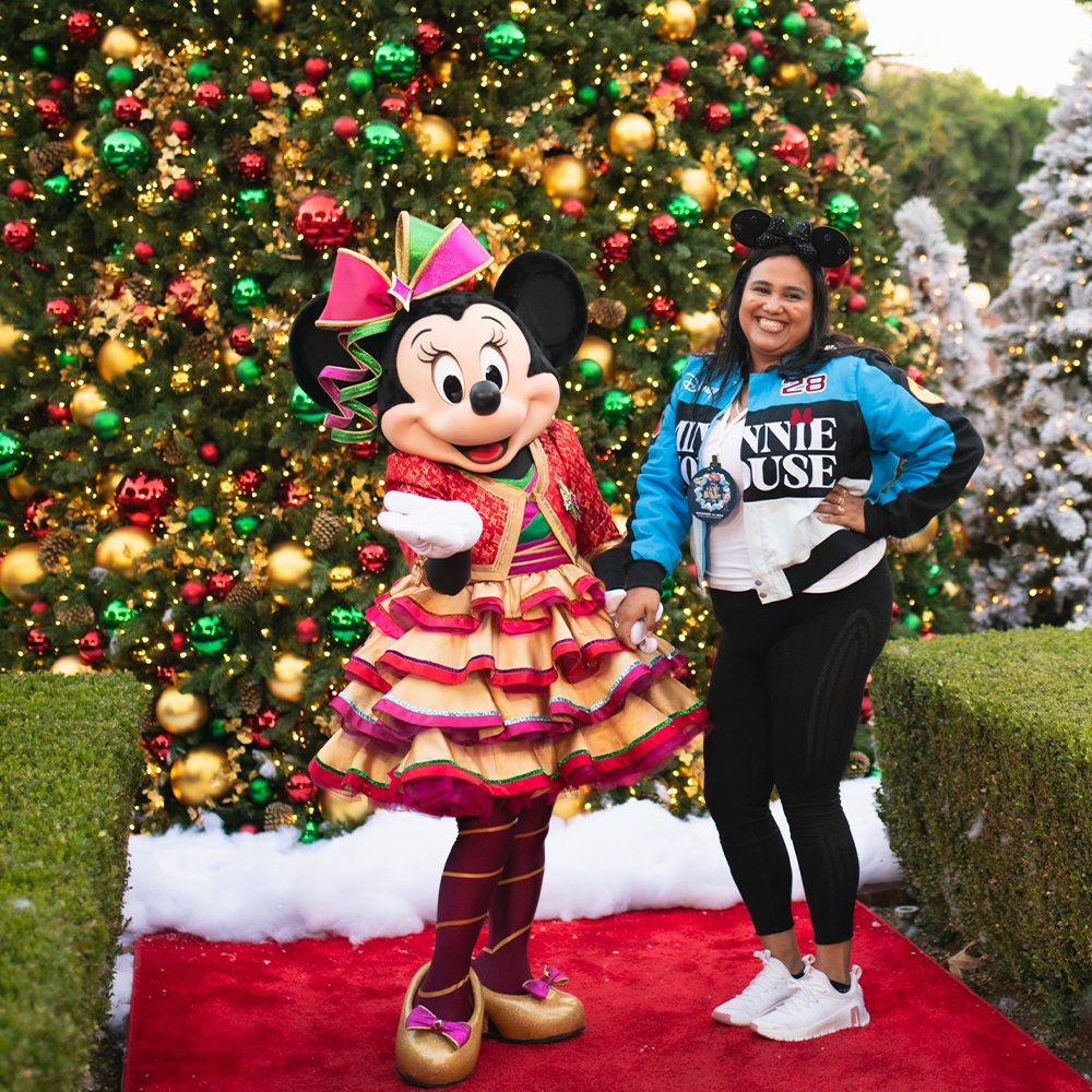 A smiling guest poses with Minnie Mouse in a festive holiday outfit on a red carpet in front of a large Christmas tree decorated with red, green, and gold ornaments.