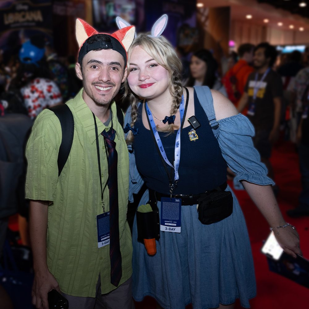 Two smiling fans wearing themed animal ear headbands pose together at a crowded convention floor, each wearing event badges and backpacks amid colorful booths and attendees in the background.
